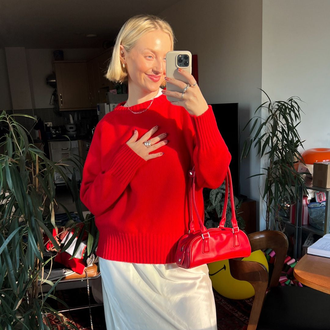 Person wearing a red sweater and white skirt holding a red handbag in a room with plants and furniture.