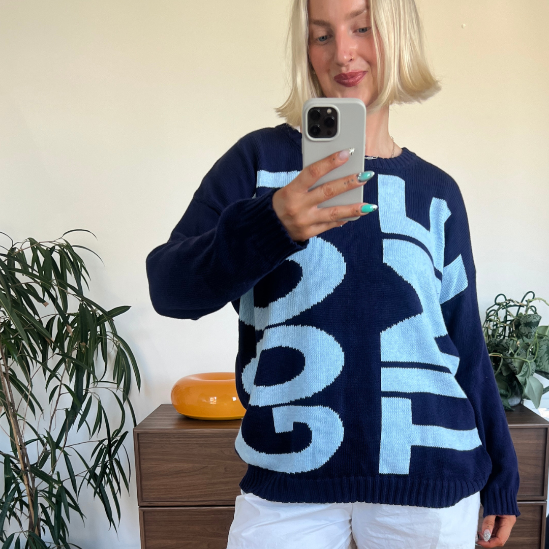 A woman takes a mirror selfie wearing a navy blue knit sweater with large light blue and white lettering, paired with white shorts, in an indoor room setting.