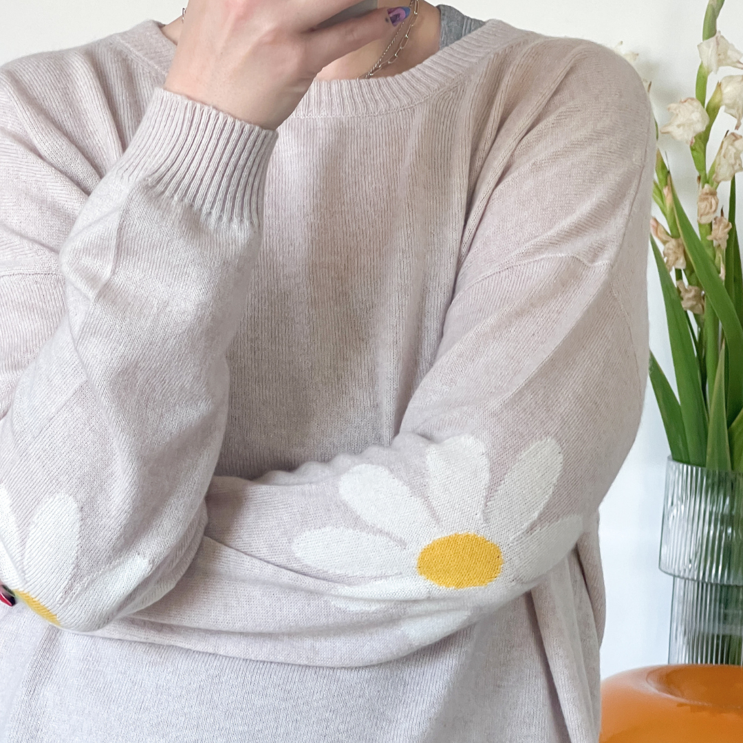 A person wearing a light beige knit sweater with a large white daisy motif and yellow center on the sleeve, posed indoors with a vase and orange object in the background.
