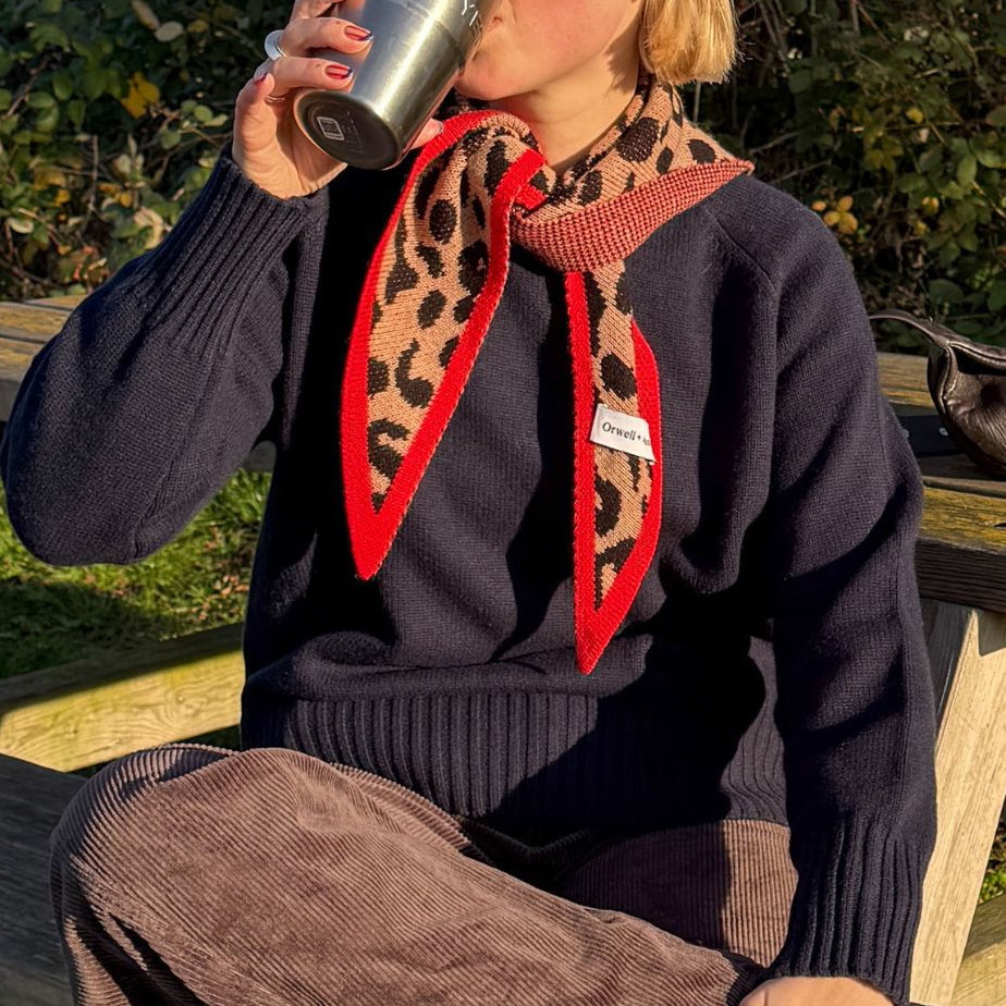 Person sitting outdoors, drinking from a metal cup, wearing a dark sweater with a red and orange pattern.