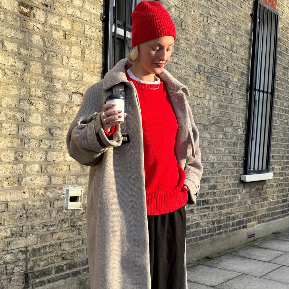 Person wearing a red beanie and sweater with a long coat, standing against a brick wall.