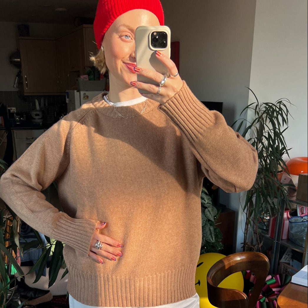 Person wearing a camel coloured sweater and red beanie taking a mirror selfie in a casual indoor setting.