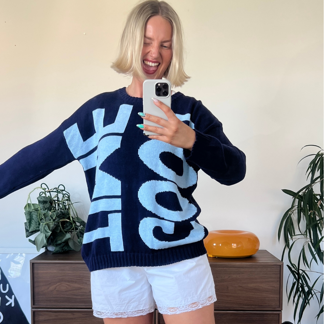 A woman takes a mirror selfie wearing a navy blue knit sweater with large light blue and white lettering that reads  'GOOD TIME', paired with white shorts.