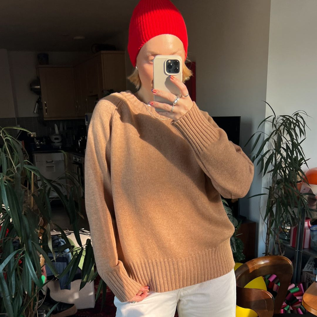 Person wearing a red beanie and a camel sweater in an indoor setting with plants and furniture.