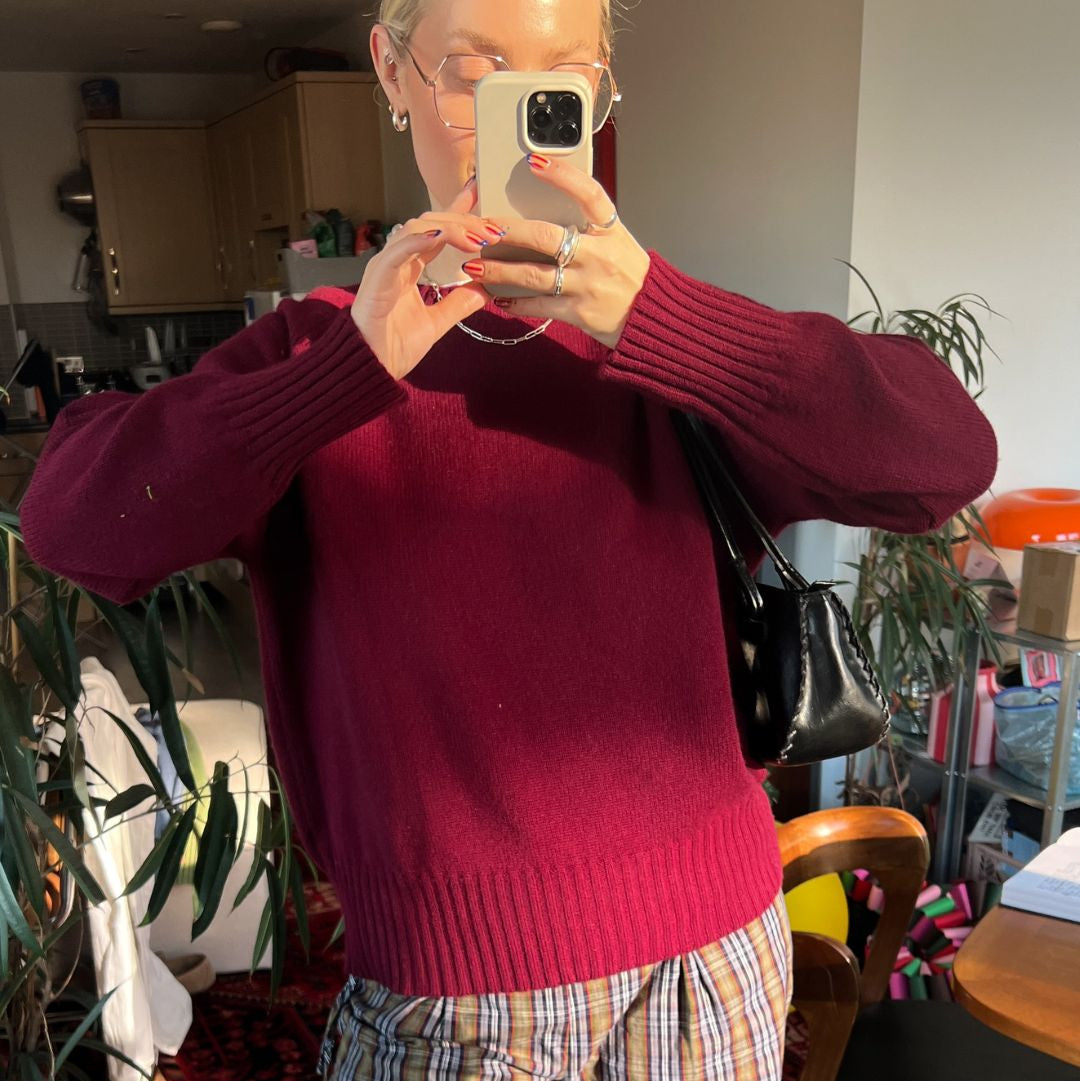 Person wearing a burgundy sweater and plaid pants taking a mirror selfie in a room with plants and furniture.
