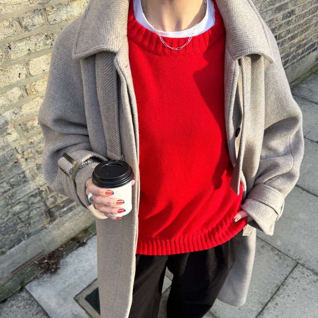 Person wearing a red sweater and beige coat holding a coffee cup on a sidewalk.
