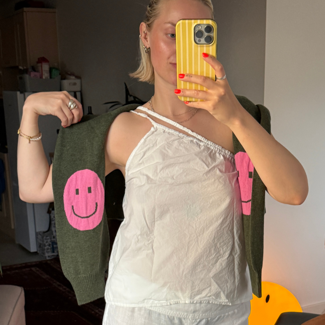Person wearing a white top and pants with a green jacket featuring pink smiley faces, taking a mirror selfie.