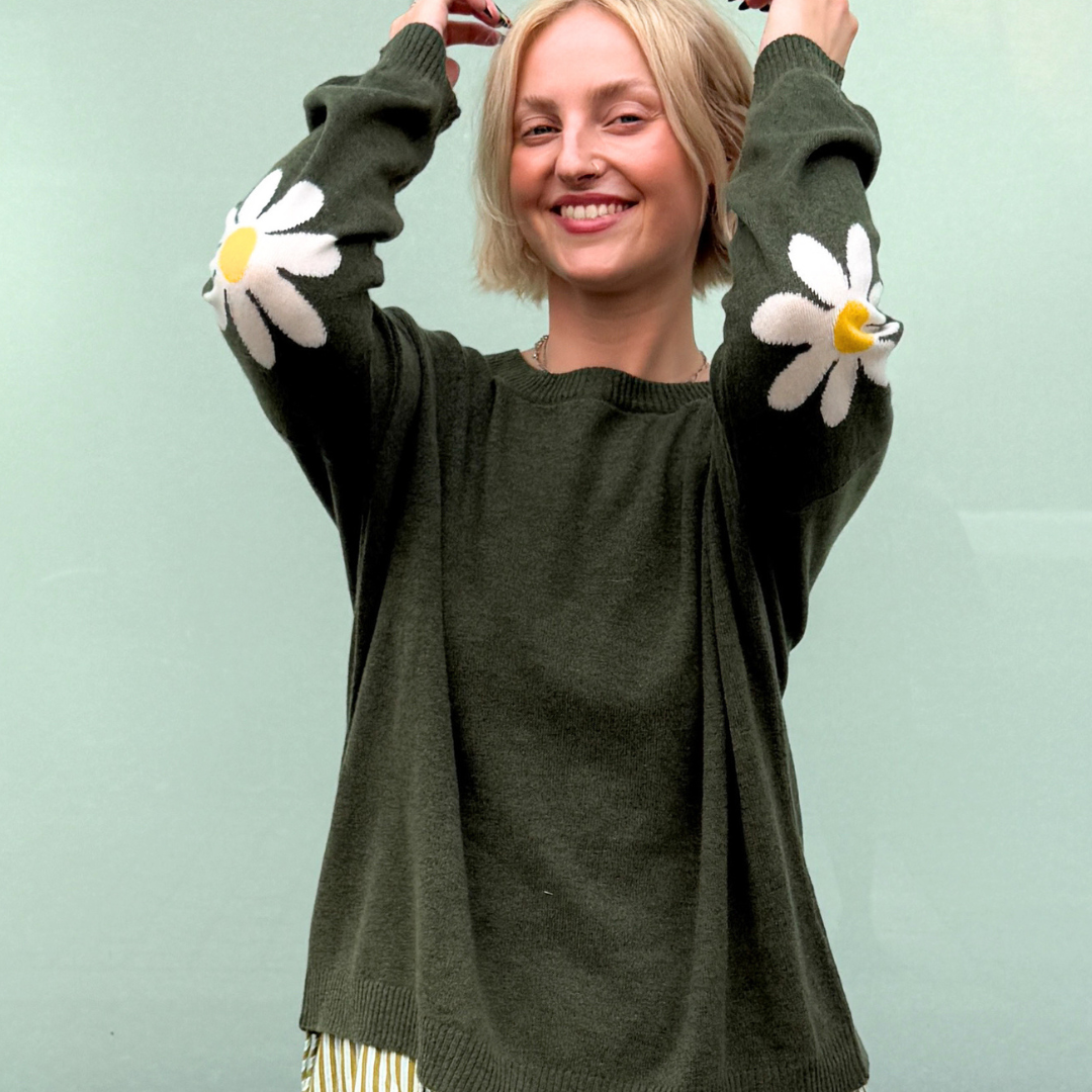 A woman wearing an olive green knit sweater with large white daisy patches on both sleeves, paired with a cream and green striped skirt, posing in front of a pale green chalkboard.